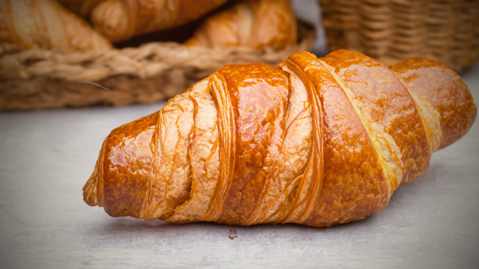 A freshly baked, golden brown croissant with flaky layers, sitting on a white plate.