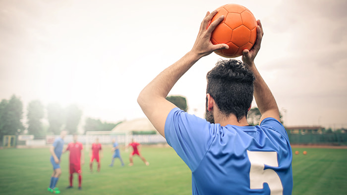 A soccer player looking confused while throwing the ball into the field, with the opposing team laughing